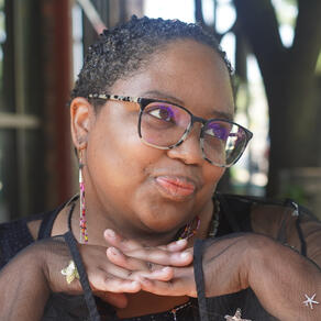 Jade, a Black person with short curly hair and tortoise-style glasses, stares off screen, holding their hands beneath their chin.